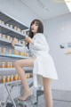 A woman in a white lab coat sitting on a stool in a pharmacy.